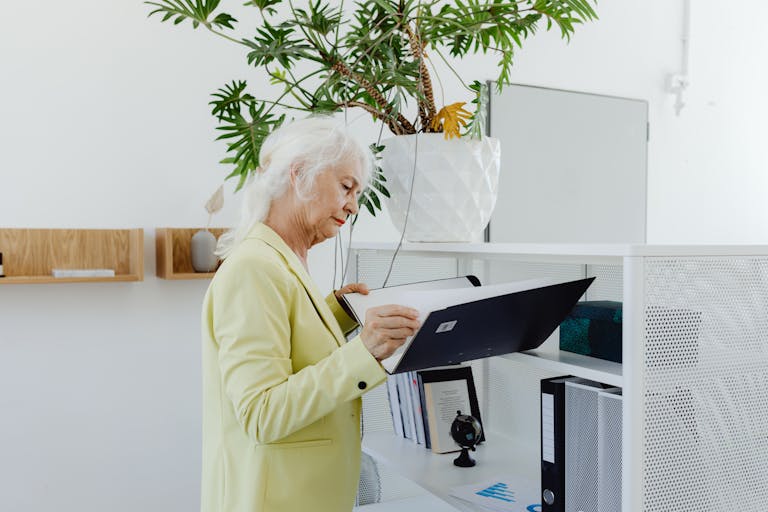 Elderly businesswoman in yellow blazer sorting through files in a modern office setting.