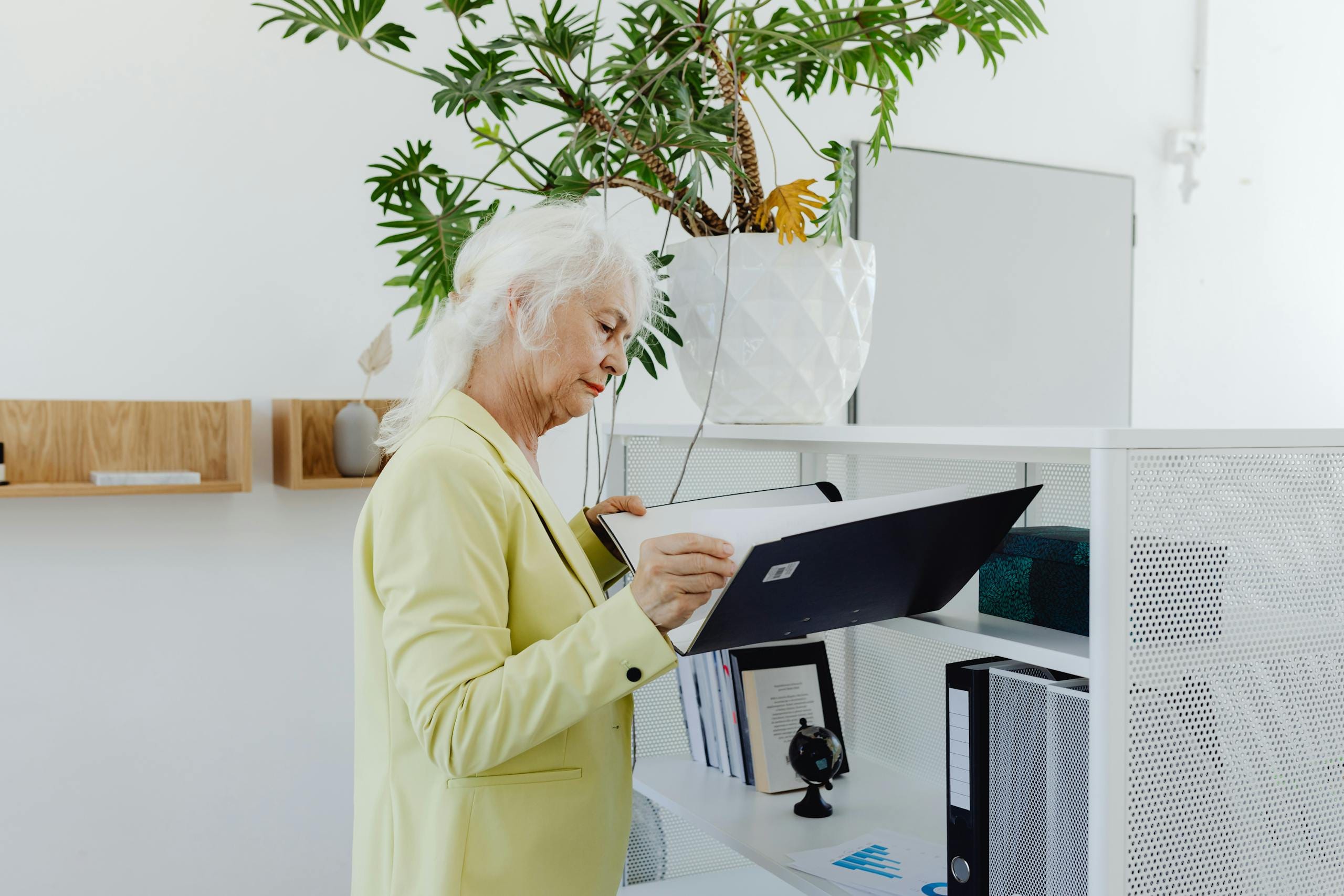 Elderly businesswoman in yellow blazer sorting through files in a modern office setting.